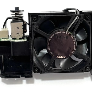 May include: A black computer fan with a square housing and four blades. The fan has a label that reads "Nidec" and "Made in China". The fan is attached to a black plastic component with a spring and electrical connectors.