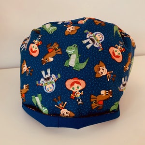 May include: A blue fabric scrub hat with a pattern of Toy Story characters, including Woody, Buzz Lightyear, and Rex. The hat has a blue trim around the bottom.