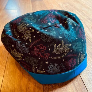 May include: A black beanie featuring colorful illustrations of the Hogwarts houses' crests and names. The hat has a blue brim and is made of a soft fabric. The crests are in gold, red, blue, and green.