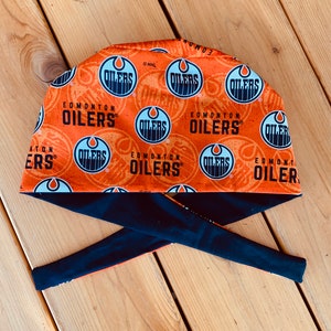 May include: An orange and black fabric headband with a repeated pattern of the Edmonton Oilers NHL logo. The headband is tied in a knot at the back.