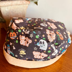 May include: A black Halloween-themed hat with a beige brim. The hat features a pattern of ghosts, pumpkins, bones, and the text "Boo-Boo Crew" and "Full Moon".
