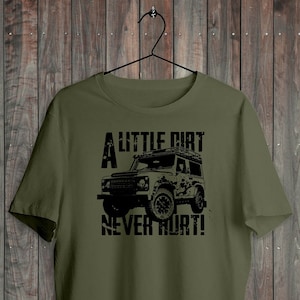 May include: Khaki green t-shirt featuring a black illustration of a 4x4 and the slogan "A LITTLE DIRT NEVER HURT!". The tee is displayed on a black hanger against a wooden backdrop. The words "DEAD SET STUDIO" are at the bottom.