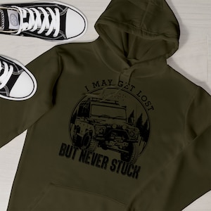 May include: An olive green hoodie featuring a black graphic of an off-road vehicle and the text "I MAY GET LOST BUT NEVER STUCK." Black and white canvas trainers are in the background.