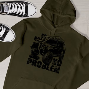 May include: An olive green hoodie featuring a black graphic of a 4x4 vehicle and the slogan "NO ROAD NO PROBLEM." A pair of black and white trainers are in the background. The hoodie has a hood and a front pocket.