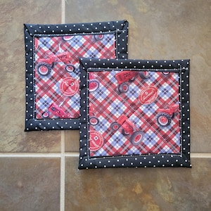 Puede incluir: Dos salvamanteles cuadrados acolchados. La tela central presenta un estampado de cuadros rojos, blancos y azules con tractores rojos y el texto "Genuine Farmall Parts". El borde es negro con lunares blancos.