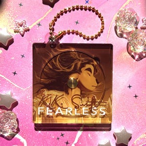 May include: A gold keychain featuring a clear acrylic charm with a gold-toned image of a woman with long hair and the text "Fearless" on it.