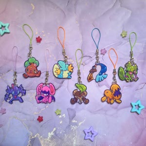 Puede incluir: Una colección de ocho coloridos dijes de acrílico con varios diseños de criaturas de dibujos animados, cada uno unido a un pequeño lazo de cordón de color. Los dijes muestran formas diversas y tonos vibrantes como morado, rojo, azul, naranja y verde.