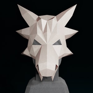 May include: A white paper mache mask shaped like a dragon's head with a pointed snout and large ears. The mask has a geometric design and is mounted on a black stand.