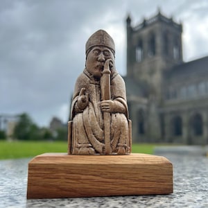 May include: A carved, beige-colored chess piece depicting a seated bishop figure, holding a staff. The figurine is mounted on a rectangular wooden base. The background features a blurred stone building under an overcast sky.