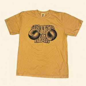 May include: Mustard yellow t-shirt with a black graphic. The graphic features the words "Grateful Dead" in a stylized font, with two circular designs on either side. The shirt is made of a soft, comfortable material.