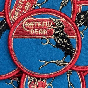 May include: Embroidered patches featuring a black crow perched on a branch within a red and blue circular design. The text "Grateful Dead" is arched above the crow. The patches are on a blue fabric background.