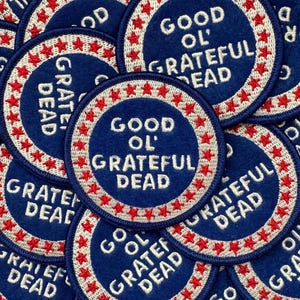 May include: A pile of blue circular patches, each embroidered with the words "GOOD OL' GRATEFUL DEAD" in white. The patches have a white border with red star detailing. The patches are overlapping.