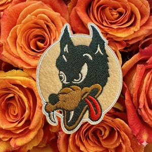 May include: A circular embroidered patch featuring a stylized black wolf head with a tan snout, white fangs, and a bright red tongue. The design is set against a light beige felt background with a white stitched border, showcasing detailed thread work.