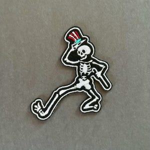 May include: An embroidered patch featuring a white skeleton in a dynamic walking pose, wearing a top hat with red, white, and blue stripes. The skeleton is outlined in black thread, with one arm raised to the hat and the other arm extended outward.