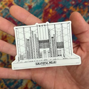 May include: A white sticker featuring a black and white illustration of a stage setup. The sticker includes the text "Grateful Dead" and "Hollywood Bowl - July 1974". The design depicts speakers, lights, and other stage elements.