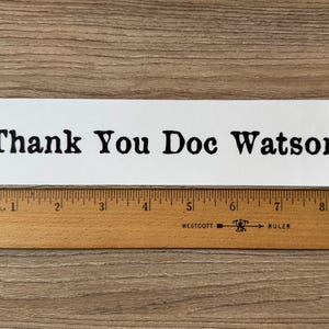 May include: A white rectangular sign with the text "Thank You Doc Watson" in black, placed on a wooden surface. A wooden ruler with inch markings is below the sign, measuring up to 9 inches.