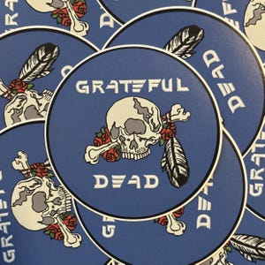 May include: A collection of round blue stickers featuring a skull with crossbones, roses, and a feather. The words "Grateful Dead" are printed in white. The stickers have a white border.