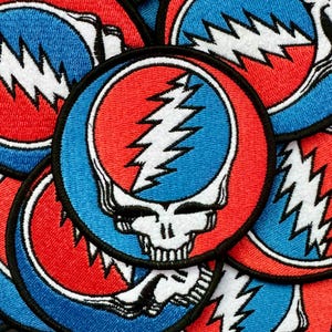 May include: A collection of embroidered patches featuring a stylized skull with a lightning bolt. The patches have a circular design with a red and blue color scheme, and a black border. The skull is white.