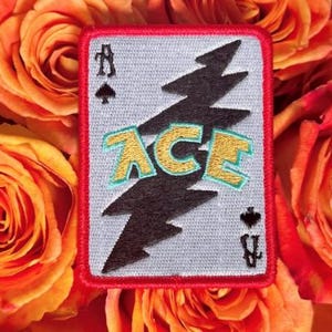 May include: A rectangular embroidered patch featuring a light grey background with a red border. A large black lightning bolt runs diagonally across the centre. The word ACE is stitched in gold with a teal outline. Black spade symbols appear in the corners.
