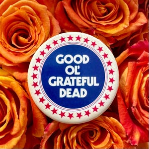 May include: A circular badge with the words "GOOD OL' GRATEFUL DEAD" in white on a blue disc, encircled by red stars. The badge is placed on a bed of orange and red roses.