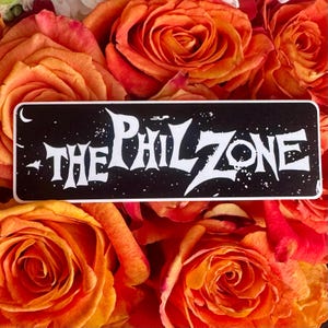 May include: A rectangular sticker with the text "THE PHIL ZONE" in white, stylized lettering against a black background. The sticker is surrounded by orange and red roses.