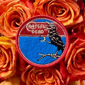 Peut inclure: Un écusson rond avec l'inscription "Grateful Dead" en blanc, représentant un corbeau noir perché sur une branche sur fond bleu et rouge. L'écusson est posé sur des roses orange.