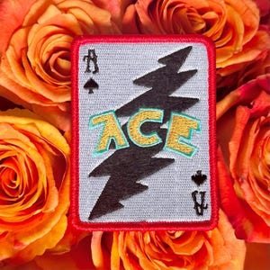May include: Embroidered patch featuring a playing card design with the word "ACE" in gold and teal lettering. The patch has a black lightning bolt design and the ace of spades and clubs symbols. The patch is framed with a red border.