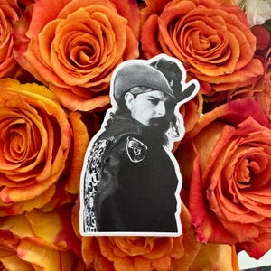 May include: A black and white sticker featuring a portrait of a man in a cowboy hat and jacket. The jacket has a decorative design on the back. The sticker is set against a backdrop of orange roses.