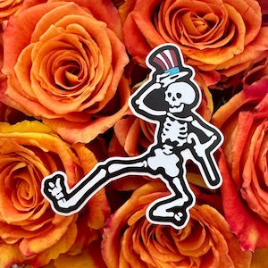 May include: A black and white sticker featuring a dancing skeleton wearing a top hat with red and blue stripes. The sticker is set against a backdrop of orange roses.