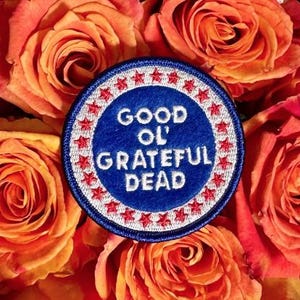 Peut inclure: Un écusson brodé circulaire avec un centre bleu profond portant le texte blanc GOOD OL' GRATEFUL DEAD. La bordure est un anneau blanc décoré de petites étoiles rouges, entouré d'un fin liseré bleu, posé sur des roses orange et roses.