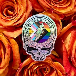 May include: A holographic sticker featuring a skull design with a colorful tree and rainbow accents. The sticker has the text "Live Your Life Praised By Its Own Design" around the edge. The sticker is set against a background of orange roses.