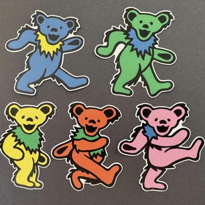 May include: Five die-cut stickers featuring stylised dancing bears in blue, green, yellow, orange, and pink. Each bear has a contrasting coloured neck ruff, black facial features, and a thick black outline, presented as a set of individual graphic decals.