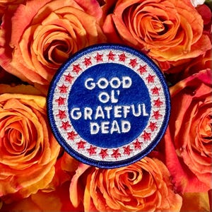 Peut inclure: Un écusson rond bleu avec l'inscription "GOOD OL' GRATEFUL DEAD" en blanc, entouré d'étoiles rouges. L'écusson est posé sur un fond de roses orange et rouges.