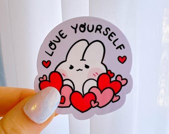 May include: A purple sticker with a white bunny surrounded by red hearts. The sticker says "LOVE YOURSELF" in a handwritten font.