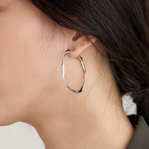 May include: Silver hoop earrings with a slightly irregular, wavy shape. The earrings are worn by a person with long dark hair and a silver chain necklace.