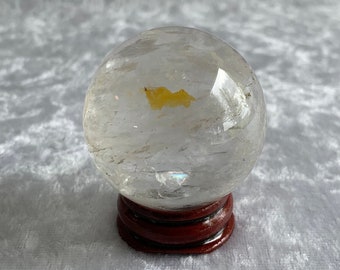 Op de afbeelding: Een heldere kwartskristallen bol met een kleine gele insluiting, die op een houten standaard staat.