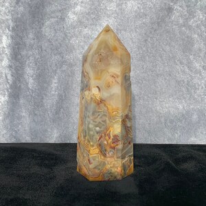 Puede incluir: Una punta de cristal de ágata marrón, blanco y gris con una parte superior puntiaguda y una base plana. El cristal está sobre una superficie negra.