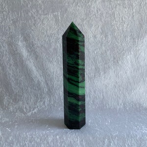 Może przedstawiać: Zielono-czarna, paskowana, szpiczasta kryształowa forma, znana również jako różdżka lub obelisk, jest prezentowana na białej, fakturowanej powierzchni.