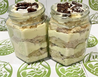 Puede incluir: Dos frascos de vidrio llenos de pastel vegano en capas. El pastel tiene una base de color claro con relleno cremoso y está cubierto con trozos de chocolate y migas blancas. Los frascos están sobre una superficie con un logotipo verde.
