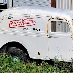 May include: A vintage white Krispy Kreme Doughnuts delivery truck, showing the side and front. The truck's logo is in red script. The vehicle is weathered, with rust visible. The truck is parked in tall grass.