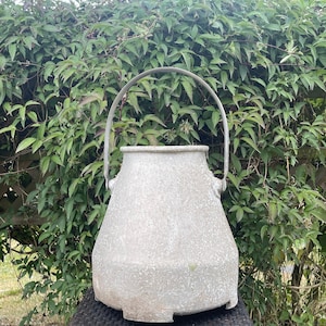 Puede incluir: Una regadera grande de metal gris con asa. La regadera tiene una superficie texturizada y un fondo redondeado.