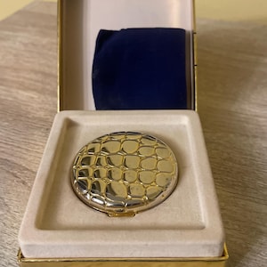 Peut inclure: Un miroir de maquillage compact doré avec un motif texturé en forme d'écailles, présenté dans un coffret ouvert bordé d'or avec un intérieur beige. Une pochette en velours bleu foncé est visible. Le compact est rond et à charnière.