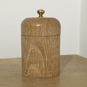 Puede incluir: Un tarro de almacenamiento de madera con un pomo de latón en la tapa. El tarro está hecho de madera marrón claro con un patrón de veta natural.
