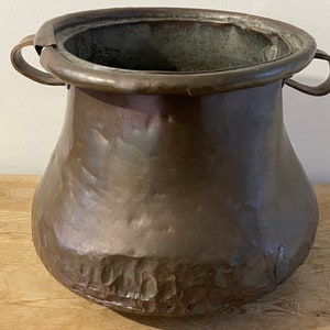 Puede incluir: Una olla de cobre antigua grande con dos asas. La olla tiene una forma redondeada y bulbosa y una textura martillada.