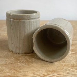 May include: Two vintage ceramic jars with a ribbed design. The jars are a light gray color and have a worn, rustic appearance.