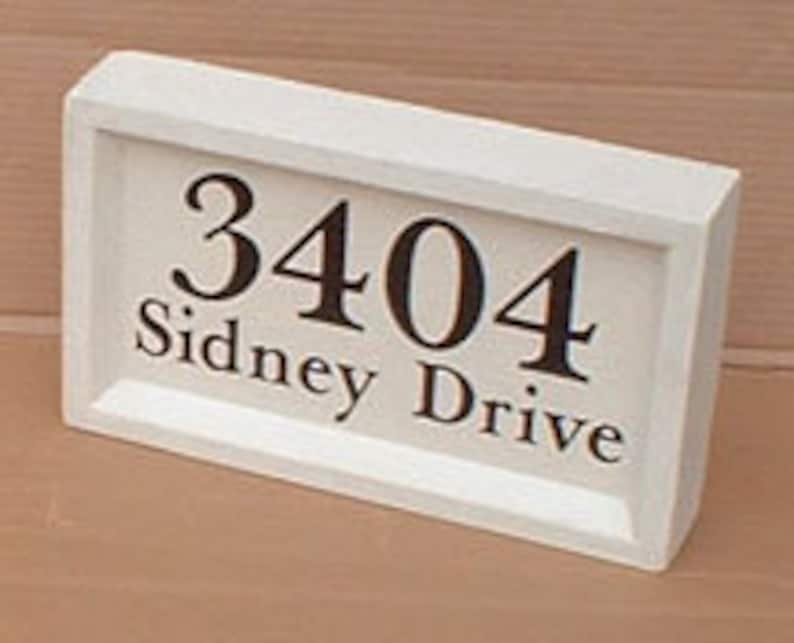 May include: A rectangular, off-white wooden address plaque. The plaque displays the numbers "3404" in a dark brown font above the words "Sidney Drive". The frame has a simple, beveled edge.