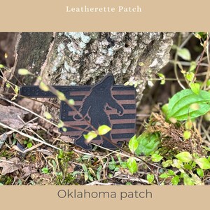 May include: A black and brown leatherette patch in the shape of Oklahoma, featuring a silhouette of Bigfoot. The patch has brown horizontal stripes and the text "Oklahoma patch" at the bottom. The text "Leatherette Patch" is at the top.