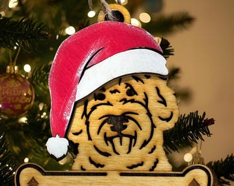 May include: Wooden Christmas ornament featuring a dog wearing a Santa hat. The ornament is light brown with a red and white hat. A bone-shaped section at the bottom has a tree design. The ornament is hanging on a Christmas tree.