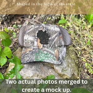 May include: Camouflage baseball cap with a Bigfoot silhouette patch. The cap has a mesh back and a curved brim. The text "Shaped in the form of the state" and "Two actual photos merged to create a mock up" are visible.