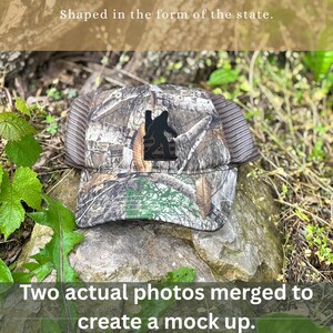 May include: A camouflage trucker hat with a black Bigfoot silhouette patch. The hat has a mesh back and a curved brim. The text "Shaped in the form of the state" and "Two actual photos merged to create a mock up" are visible.